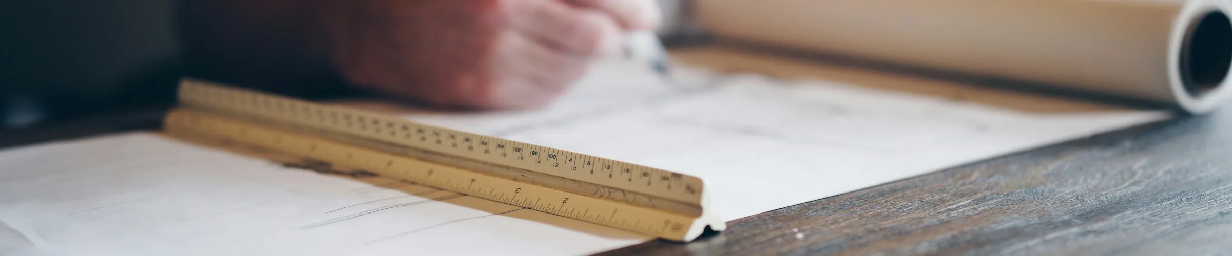 A hand drafting on a drawing sheet with a scale ruler and rolled plans
