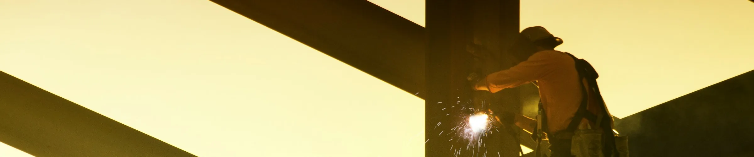 An ironworker welding at a steel beam connection, silhouetted against the sky